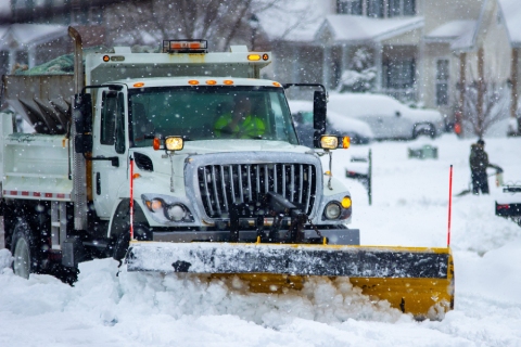 A large white dump truck with a yellow plow attachment clears a snow-covered residential street during a heavy snowfall