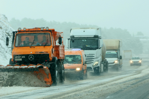 An orange Mercedes Unimog snowplow leads a line of vehicles, including semi-trucks, driving down a snow-covered highway during a winter storm