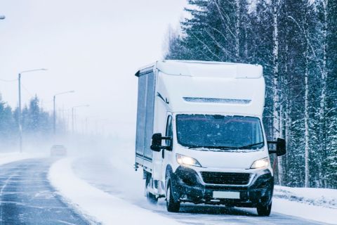 A white delivery van driving down a snow-covered road lined with trees on an overcast winter day, with its headlights illuminated