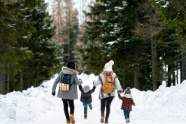 A family walks hand-in-hand down a snowy trail lined with tall evergreen trees on a winter day
