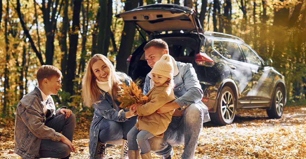 Family enjoying an autumn outing beside their SUV in a sunlit forest.