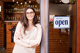Smiling woman standing with folded arms beside an “Open” sign at a storefront entrance.