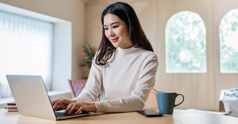 Woman working on a laptop at a table with a coffee mug and smartphone nearby.